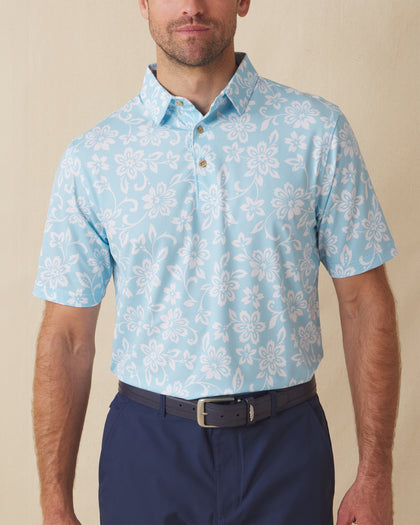 Men's Napali Coast Polo in Gemstone, front model view featuring a light aqua blue short sleeve polo with an allover white Hawaiian floral and vine print, styled with navy pants and a leather belt
