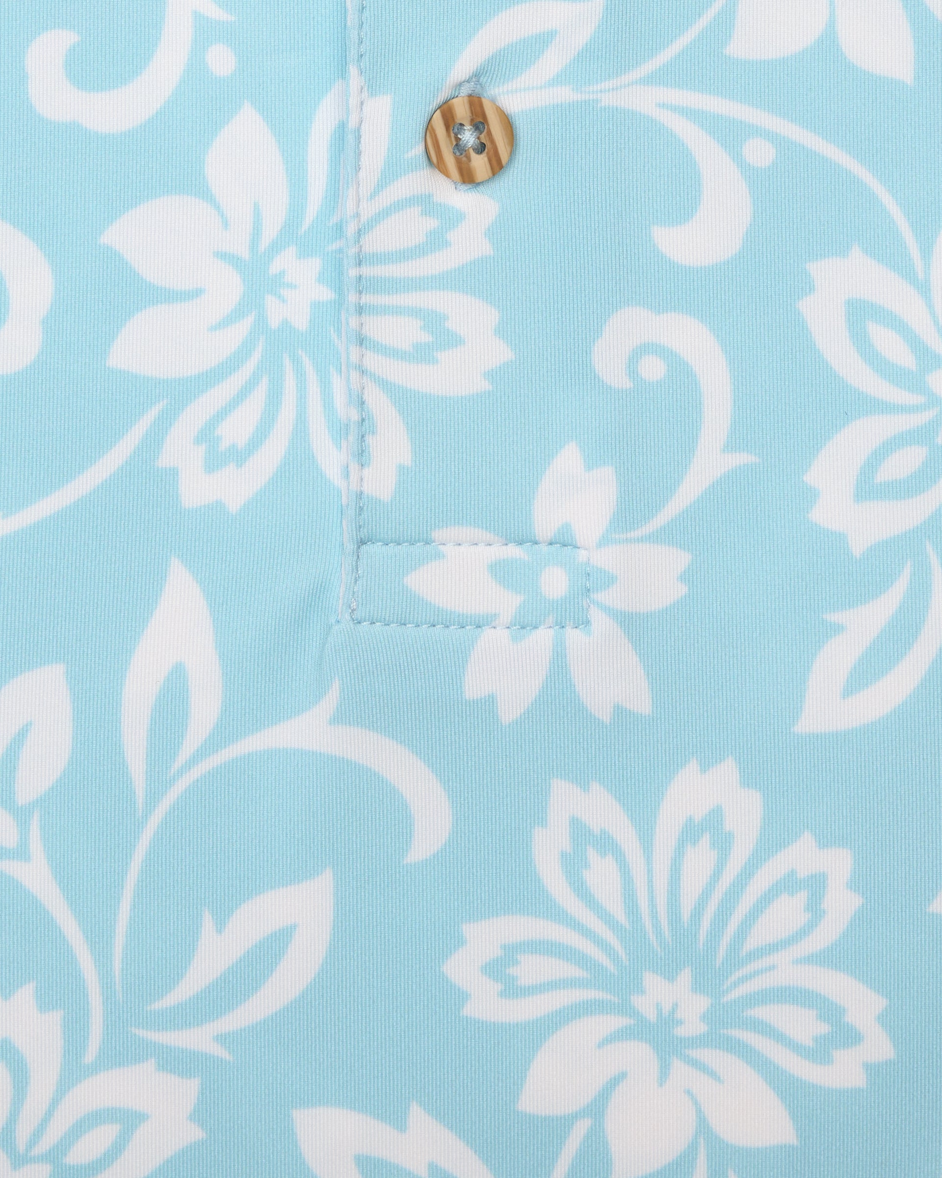 Men's Napali Coast Polo in Gemstone, close-up fabric detail view featuring an allover white Hawaiian floral and vine print on a light aqua blue performance fabric with a natural wood-tone button