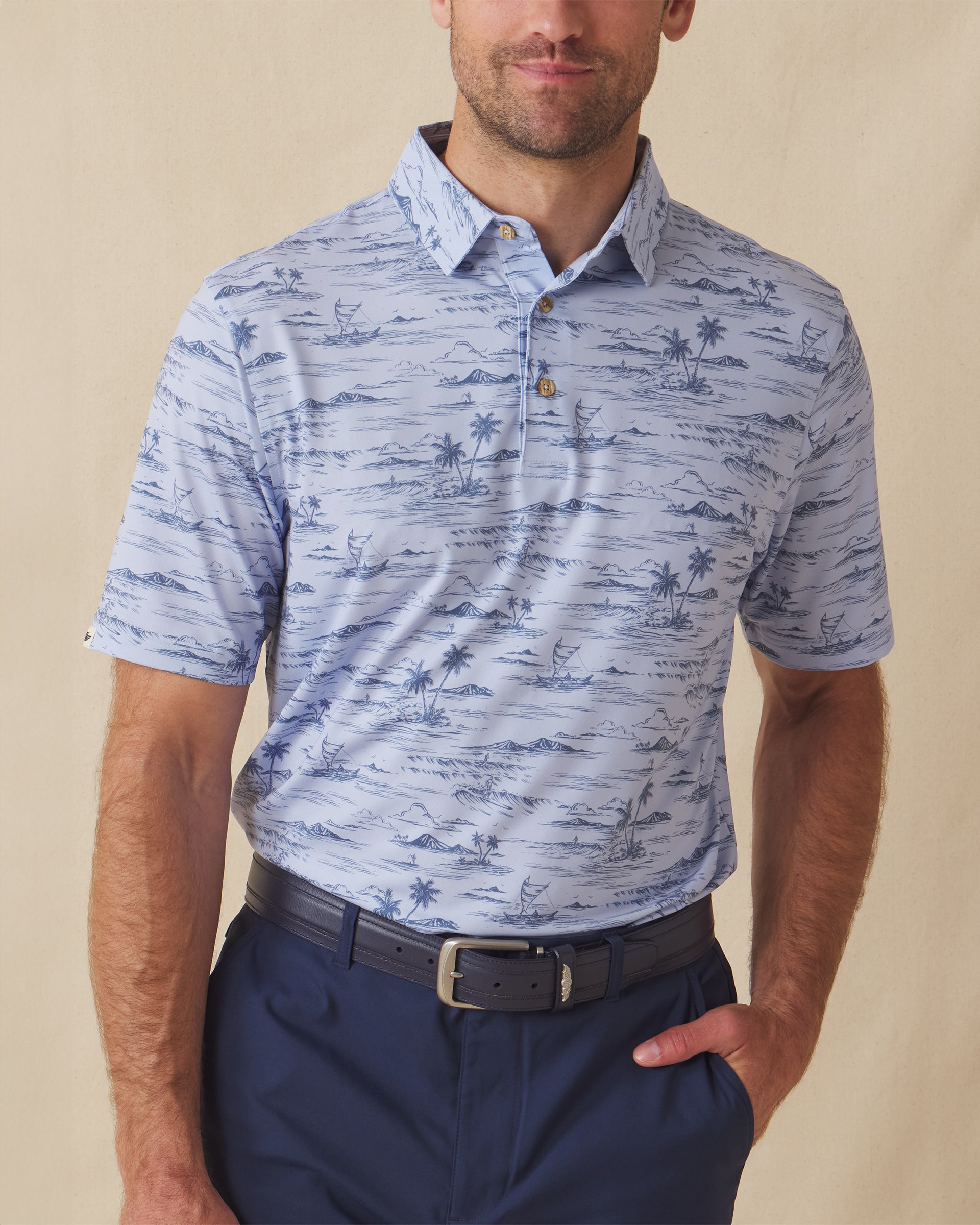 Men's Outrigger Polo in Powder, front view on model featuring a light blue Hawaiian scenic print with wood buttons, styled with navy pants and a navy belt