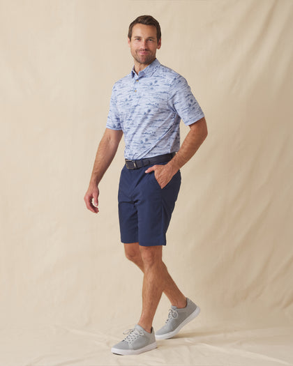 Men's Outrigger Polo in Powder, full-length side view on model featuring a light blue Hawaiian scenic print, styled with navy shorts and grey shoes