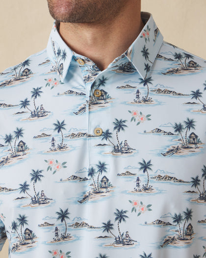 Men's Land Of Aloha Polo in Sea Spray, collar detail view featuring an allover Hawaiian island scene print with palm trees, thatched huts, hula dancers, outrigger canoes, and hibiscus flowers on a light blue performance fabric with natural wood-tone buttons