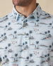 Men's Land Of Aloha Polo in Sea Spray, collar detail view featuring an allover Hawaiian island scene print with palm trees, thatched huts, hula dancers, outrigger canoes, and hibiscus flowers on a light blue performance fabric with natural wood-tone buttons