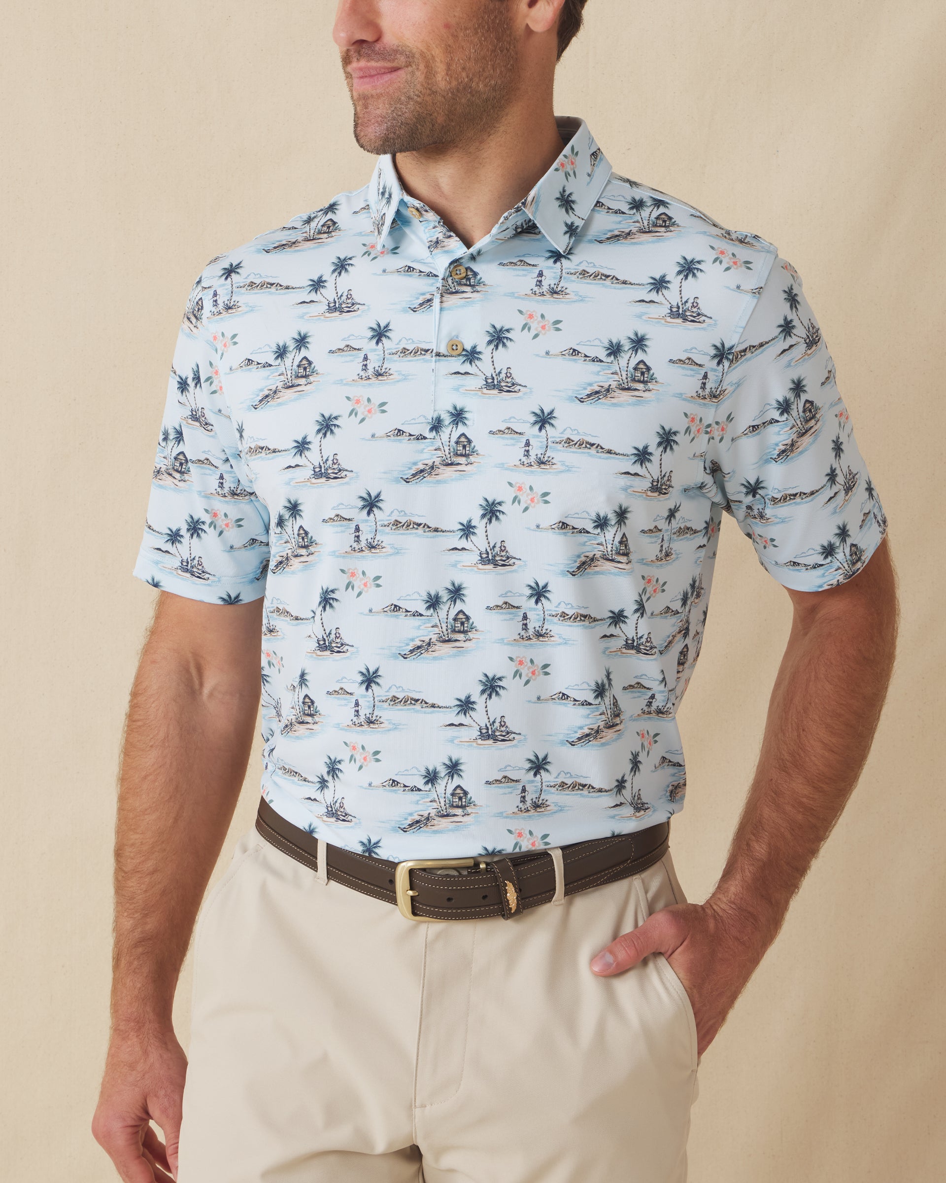 Men's Land Of Aloha Polo in Sea Spray, front model view featuring a light blue short sleeve polo with an allover Hawaiian island scene print with palm trees, thatched huts, hula dancers, outrigger canoes, and hibiscus flowers, styled with khaki pants and a leather belt