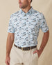 Men's Land Of Aloha Polo in Sea Spray, front model view featuring a light blue short sleeve polo with an allover Hawaiian island scene print with palm trees, thatched huts, hula dancers, outrigger canoes, and hibiscus flowers, styled with khaki pants and a leather belt