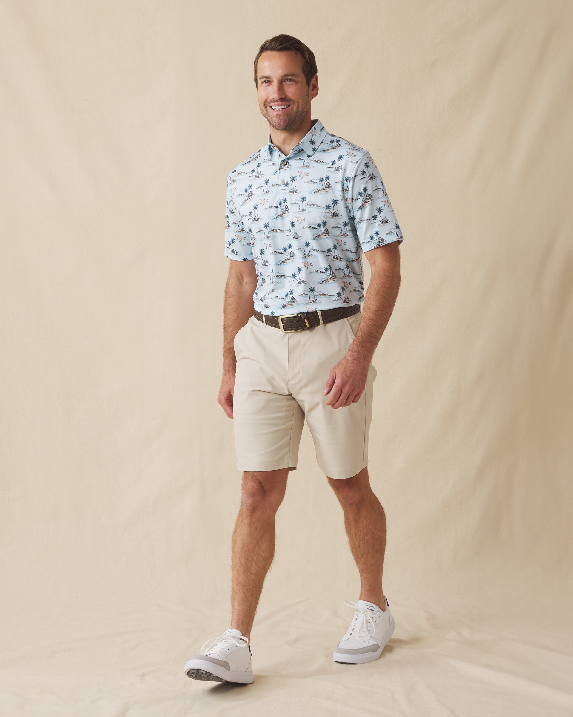 Men's Land Of Aloha Polo in Sea Spray, full length model view featuring a light blue short sleeve polo with an allover Hawaiian island scene print with palm trees, thatched huts, hula dancers, outrigger canoes, and hibiscus flowers, styled with khaki shorts and white shoes