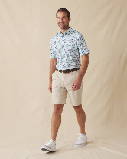 Men's Land Of Aloha Polo in Sea Spray, full length model view featuring a light blue short sleeve polo with an allover Hawaiian island scene print with palm trees, thatched huts, hula dancers, outrigger canoes, and hibiscus flowers, styled with khaki shorts and white shoes
