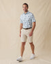Men's Land Of Aloha Polo in Sea Spray, full length model view featuring a light blue short sleeve polo with an allover Hawaiian island scene print with palm trees, thatched huts, hula dancers, outrigger canoes, and hibiscus flowers, styled with khaki shorts and white shoes