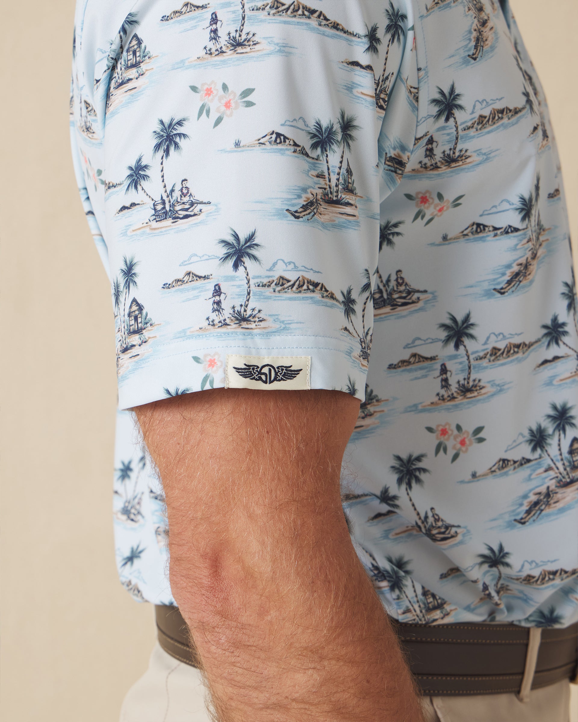Men's Land Of Aloha Polo in Sea Spray, sleeve detail view featuring a light blue short sleeve polo with an allover Hawaiian island scene print, with a Straight Down logo tab on the sleeve hem