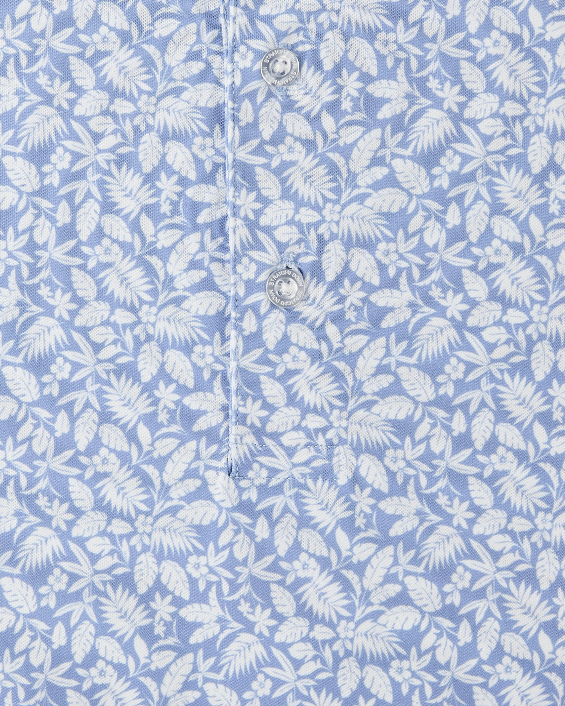 Detail view of the Tropics Lounge Polo in Blue Shade, highlighting the white botanical pattern, light blue fabric, and branded buttons.