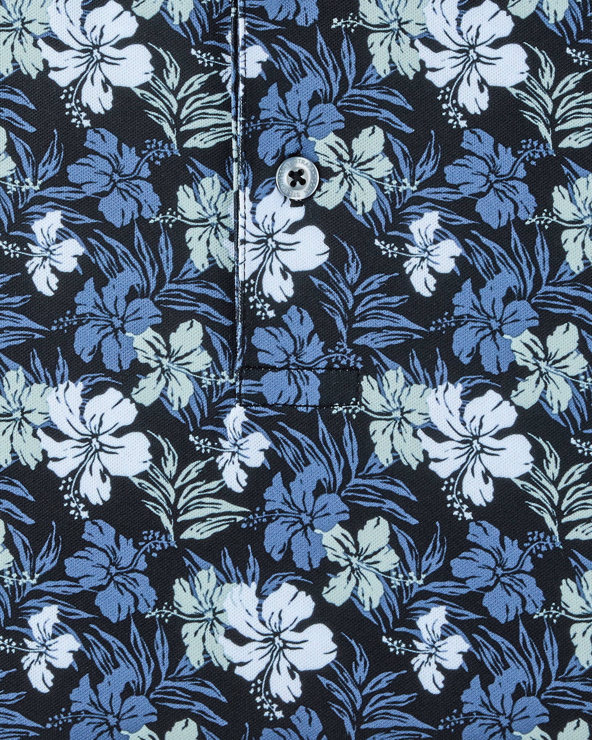 Close-up of the Hawaii 5.0 Polo showing the black fabric, blue and light green hibiscus print, button, and placket stitching.