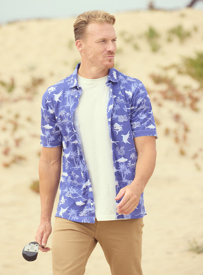 Islamorada Full Button Polo in Skipper, on model front view featuring a blue base with an all-over white nautical print of marlin, sailfish, coral, and palm trees, styled with khaki pants and worn open over a white tee on a golf course