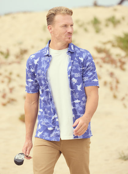 Islamorada Full Button Polo in Skipper, on model front view featuring a blue base with an all-over white nautical print of marlin, sailfish, coral, and palm trees, styled with khaki pants and worn open over a white tee on a golf course