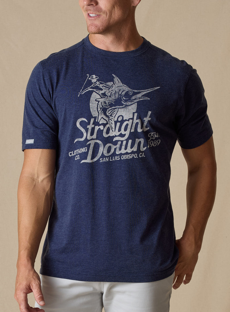 Marlin Rodeo Graphic Tee in New Indigo, front model view featuring a heathered navy short-sleeve crewneck tee with a vintage-style graphic of a golfer riding a leaping marlin, paired with light grey pants
