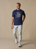 Marlin Rodeo Graphic Tee in New Indigo, full-length front model view featuring a heathered navy short-sleeve crewneck tee with a vintage-style graphic of a golfer riding a leaping marlin, paired with light grey pants and white shoes