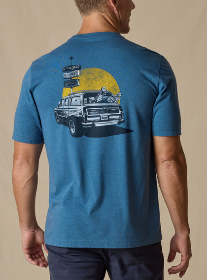 Vegas Vacation Graphic Tee in Petrol, back model view featuring a heathered blue short-sleeve crewneck tee with a vintage-style graphic of a classic woody wagon loaded with golf clubs in front of a golden sun and retro roadside, paired with navy pants