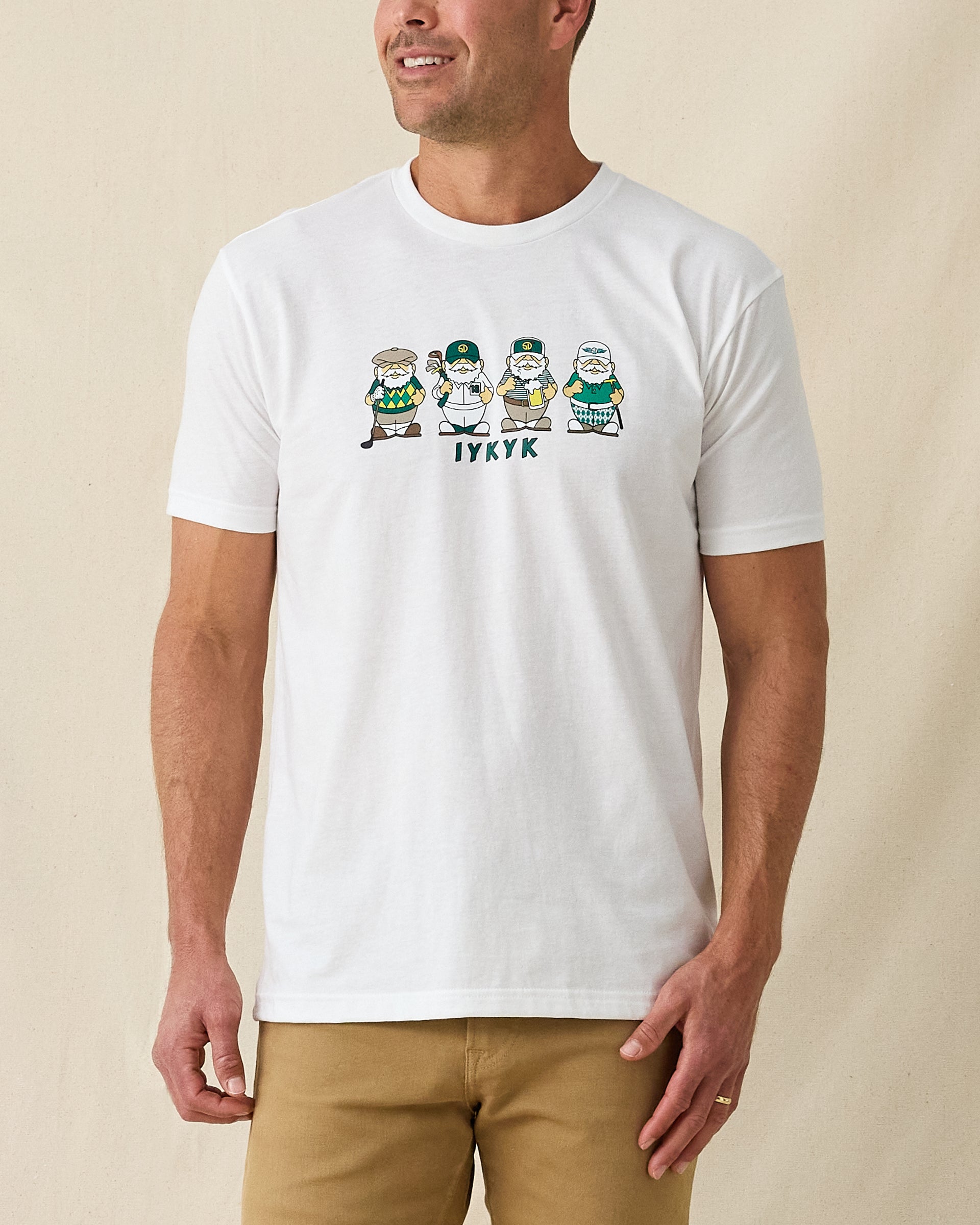 Men's IYKYK Tee in White, front view on model featuring a white base with a four golf gnome character graphic and IYKYK text in green on the chest, paired with khaki pants