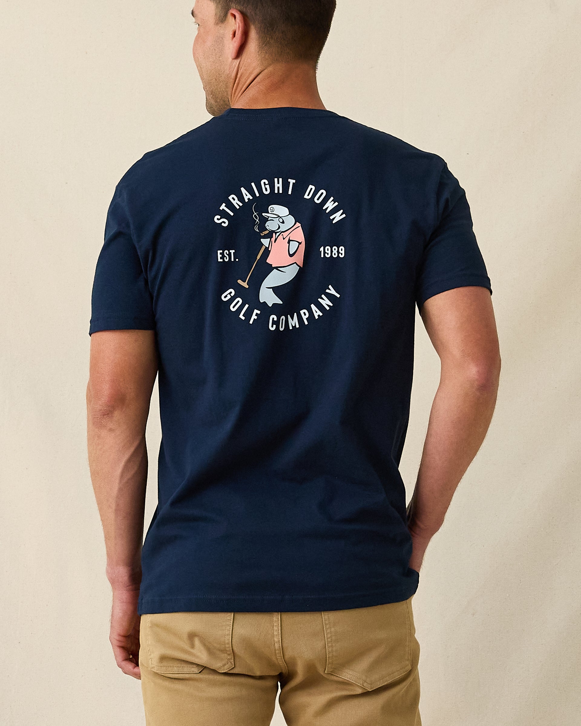 Men's Mana-Tee in Navy, back view on model featuring a large circular Straight Down Golf Company Est. 1989 graphic with cigar-smoking manatee holding a golf club, paired with khaki pants