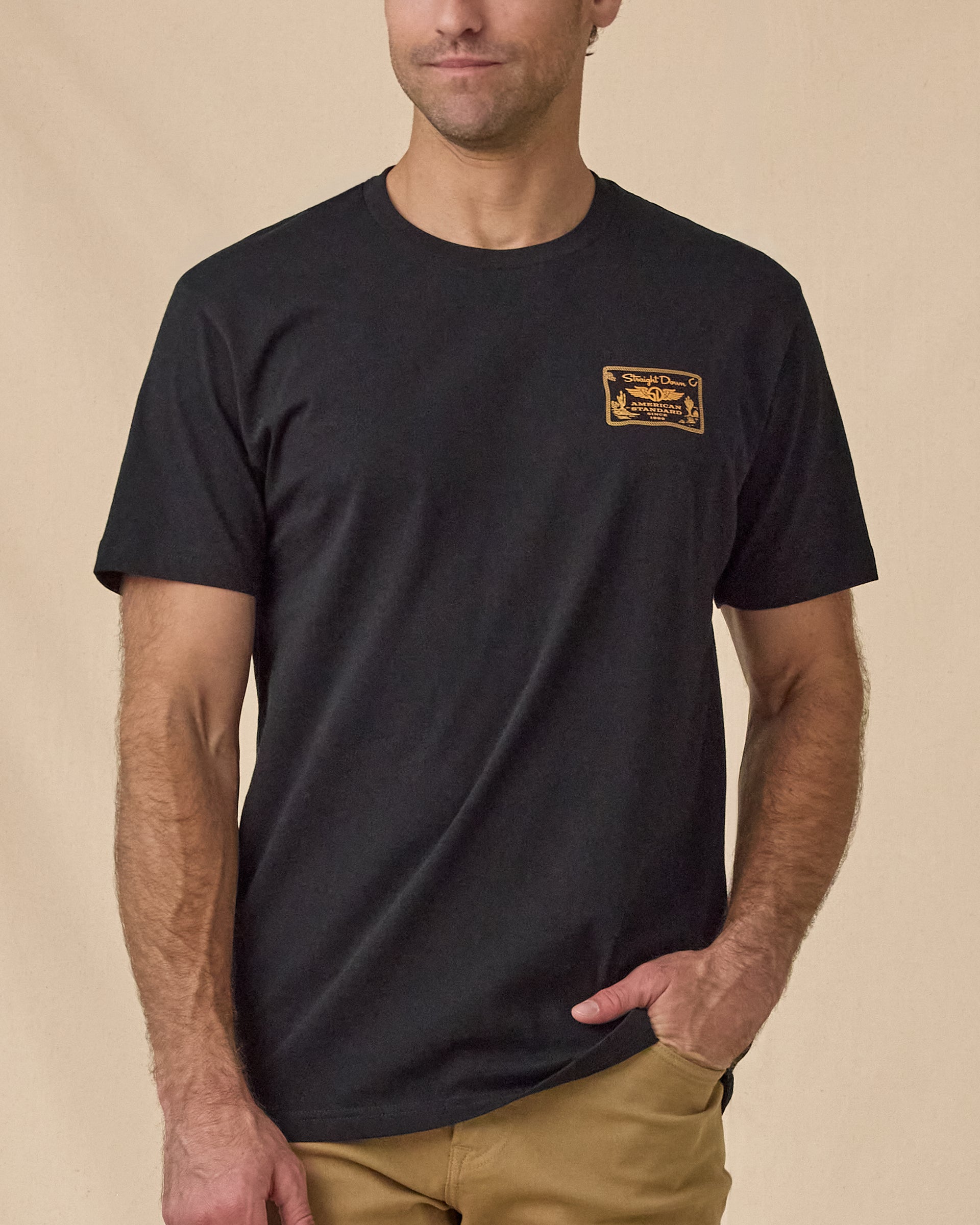 Man wearing the American Standard Tee in Black with a small gold graphic on the left chest.