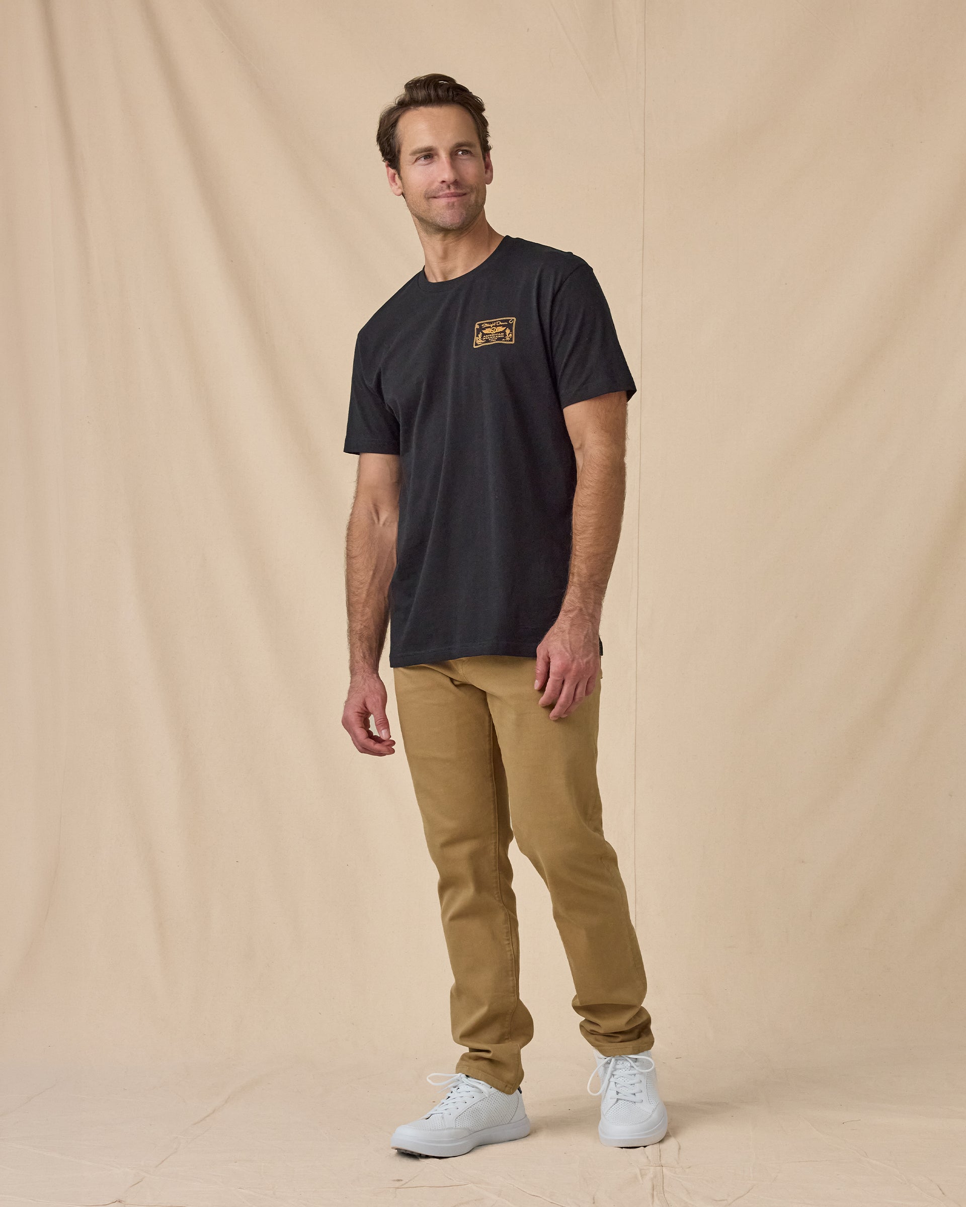 Man wearing the American Standard Tee in Black with tan pants and black sneakers.