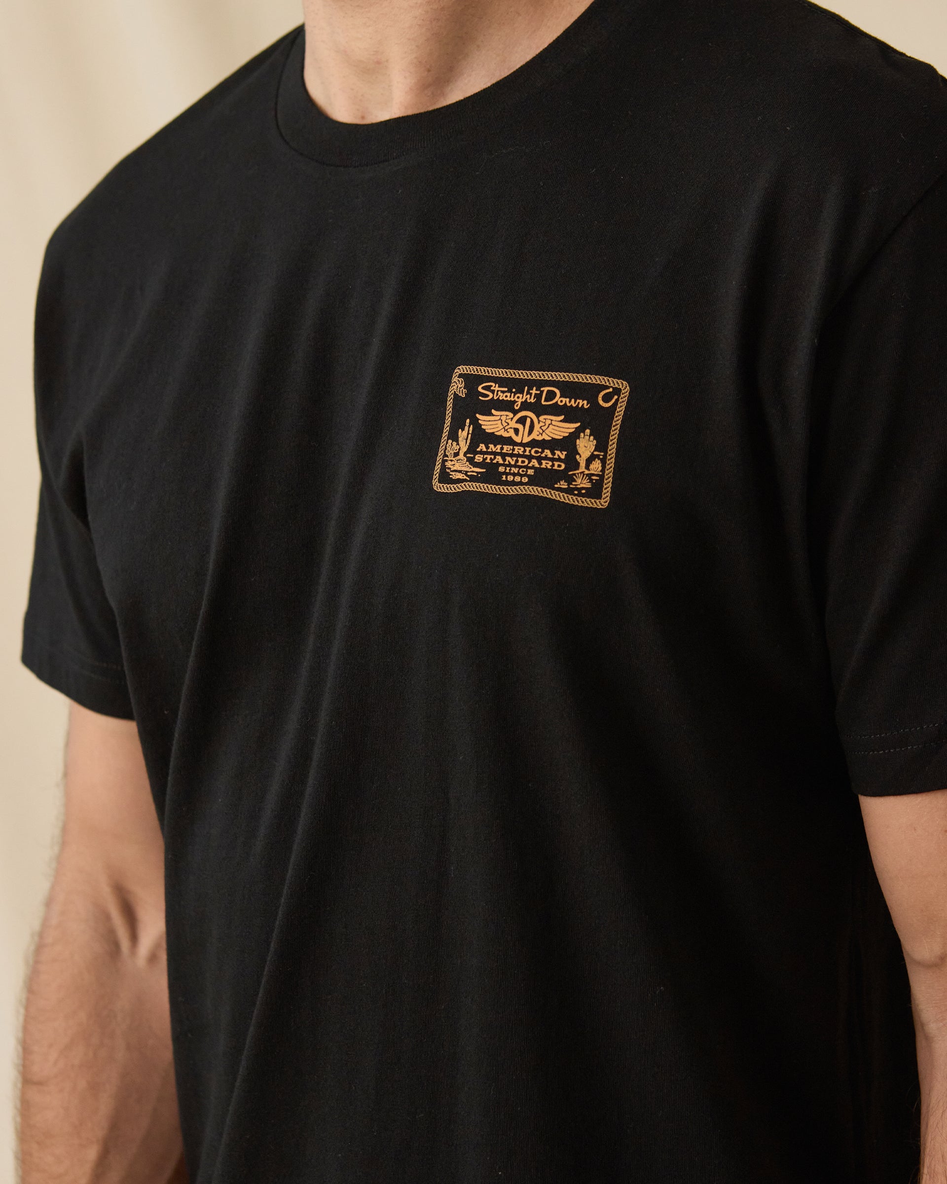 Close-up of the American Standard Tee in Black showing the small gold Straight Down American Standard graphic on the left chest.