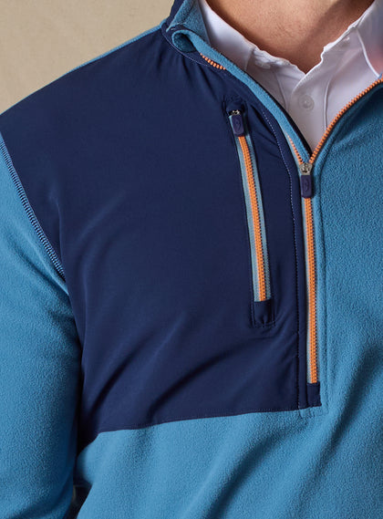 Men's Moonstone Quarter Zip in Petrol, close-up chest detail featuring navy softshell contrast panel with chest zip pocket, orange zipper tape, and branded zipper pulls layered over a white polo
