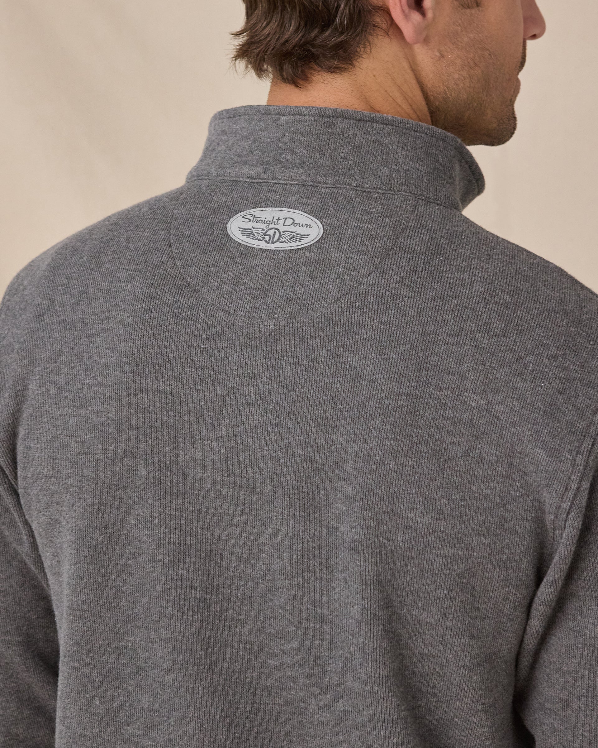 Close-up back view of a man wearing a heather gray quarter-zip pullover sweater with a small oval Straight Down logo patch on the upper neck, against a warm beige background.