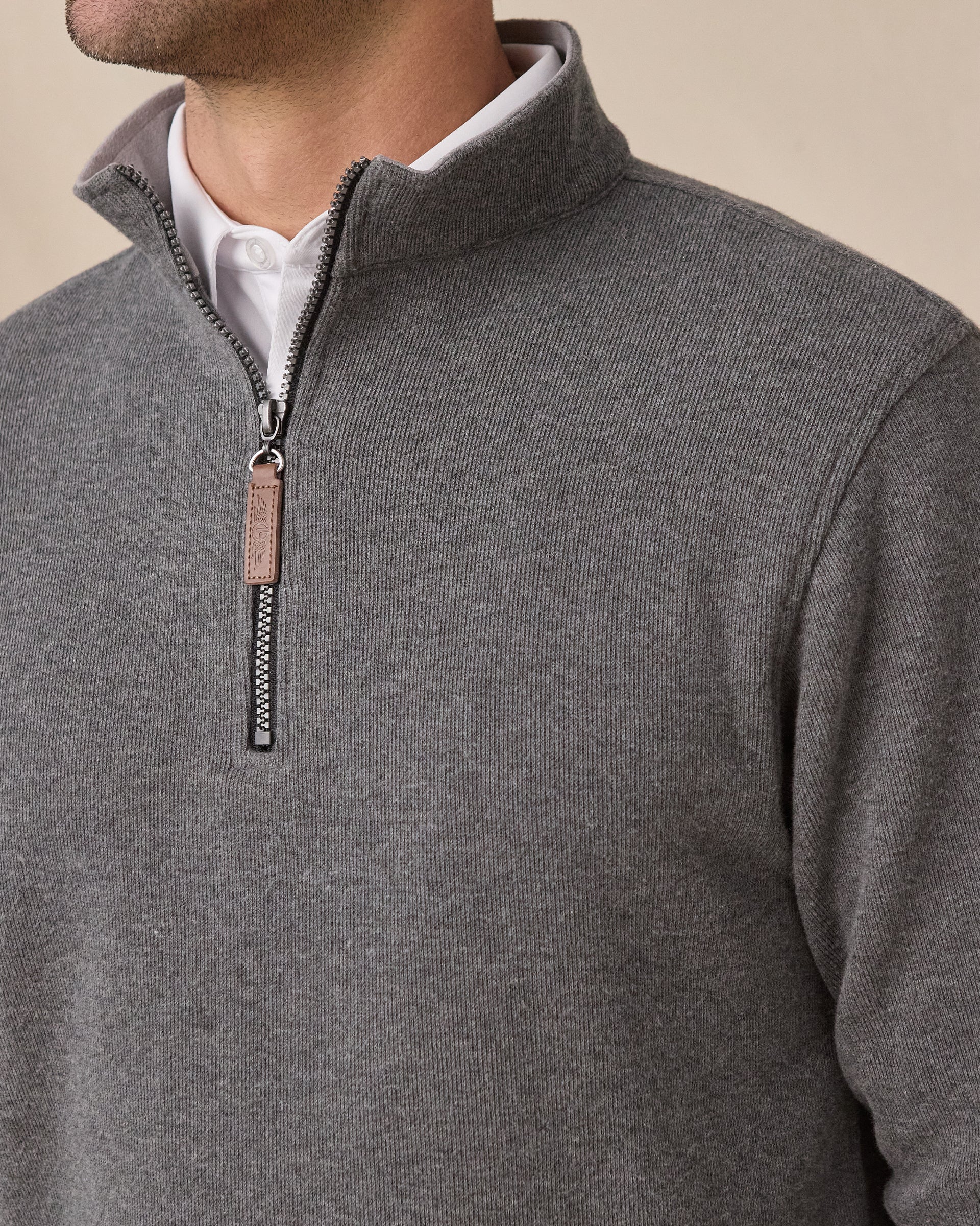 A close-up of a man wearing a heather gray quarter-zip pullover sweater over a white collared shirt, set against a plain beige background.
