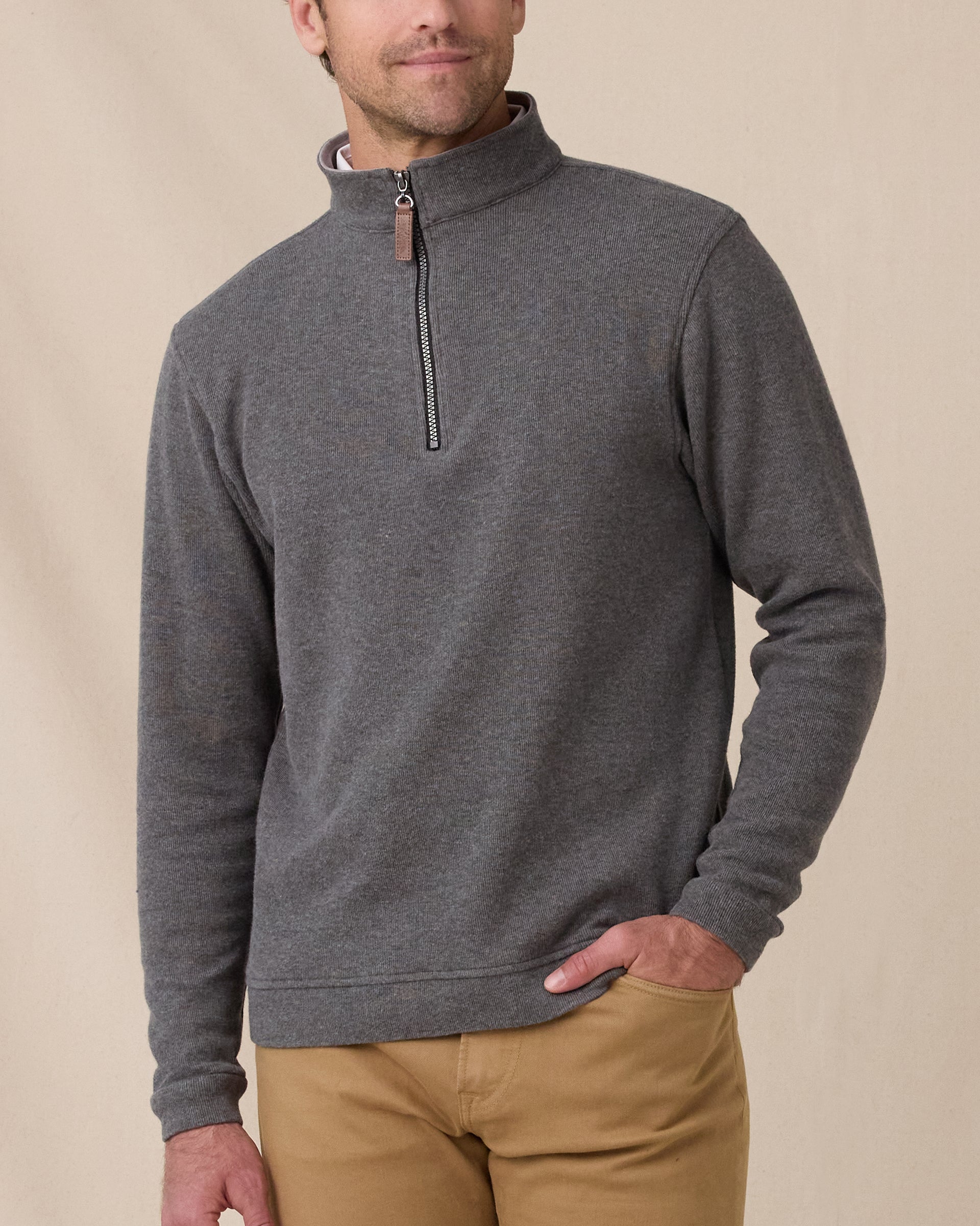 A model wearing a dark gray heathered quarter-zip pullover sweater with a brown leather zipper pull, paired with tan pants, against a neutral beige background.
