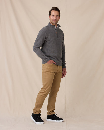 A man modeling a gray quarter-zip pullover sweater, tan pants, and black sneakers against a beige fabric backdrop.