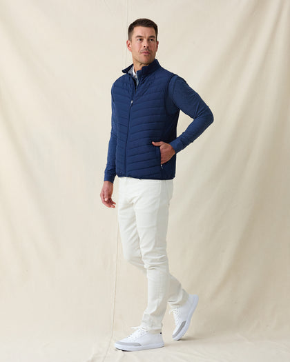 Men's Delta 2.0 Quilted Hybrid Vest in New Indigo, full length side view on model featuring a navy quilted front panel with full zip closure, layered over a navy long sleeve shirt, paired with white pants and white shoes