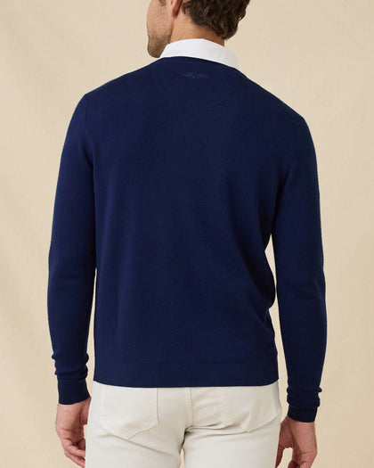 Back view of a model wearing a navy blue v-neck knit sweater over a white collared shirt and stone colored pants, photographed against a plain beige studio background.