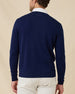 Back view of a model wearing a navy blue v-neck knit sweater over a white collared shirt and stone colored pants, photographed against a plain beige studio background.