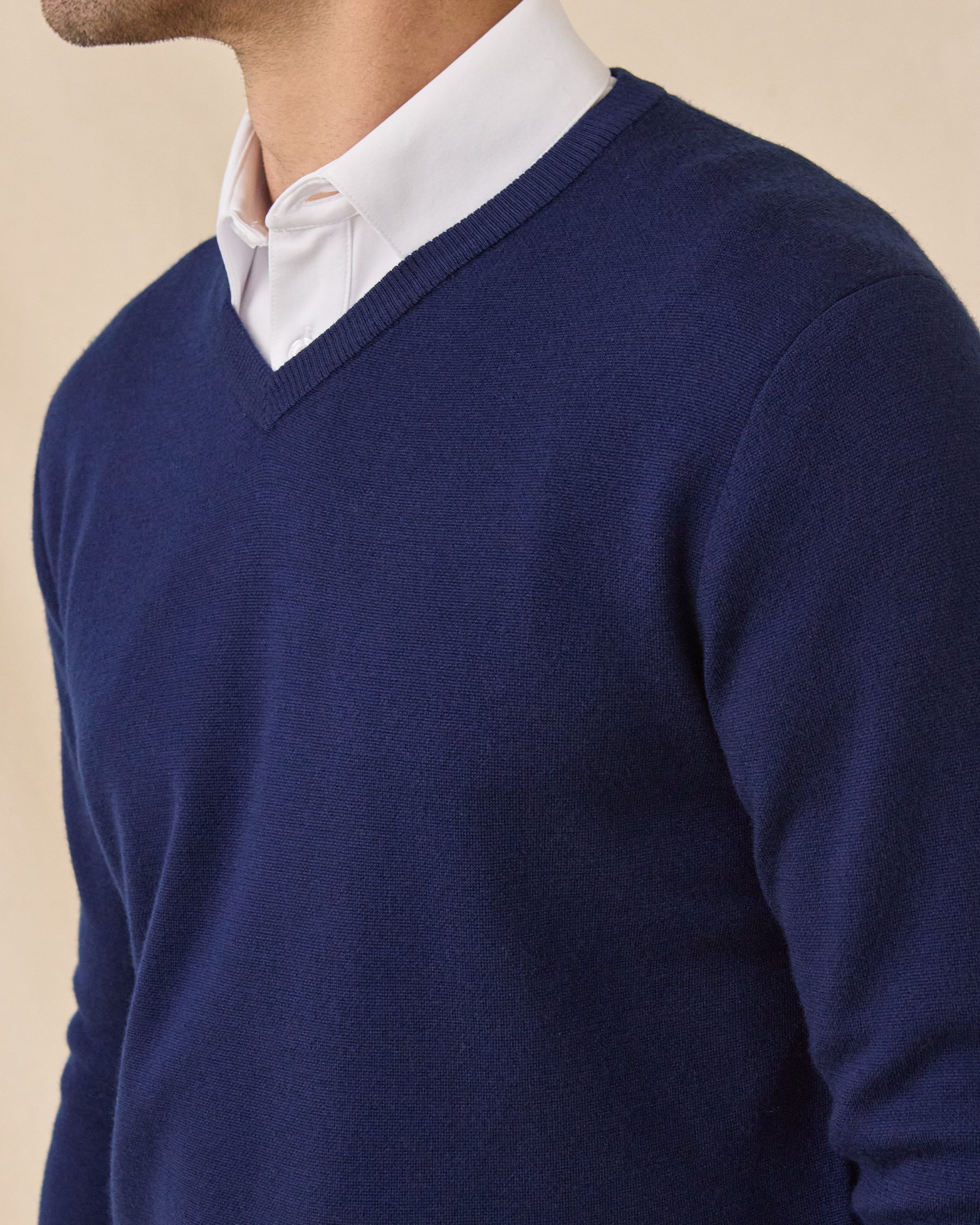 A close-up of a model wearing a navy blue V-neck knit sweater layered over a white collared shirt, against a plain beige background.
