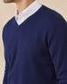 A close-up of a model wearing a navy blue V-neck knit sweater layered over a white collared shirt, against a plain beige background.