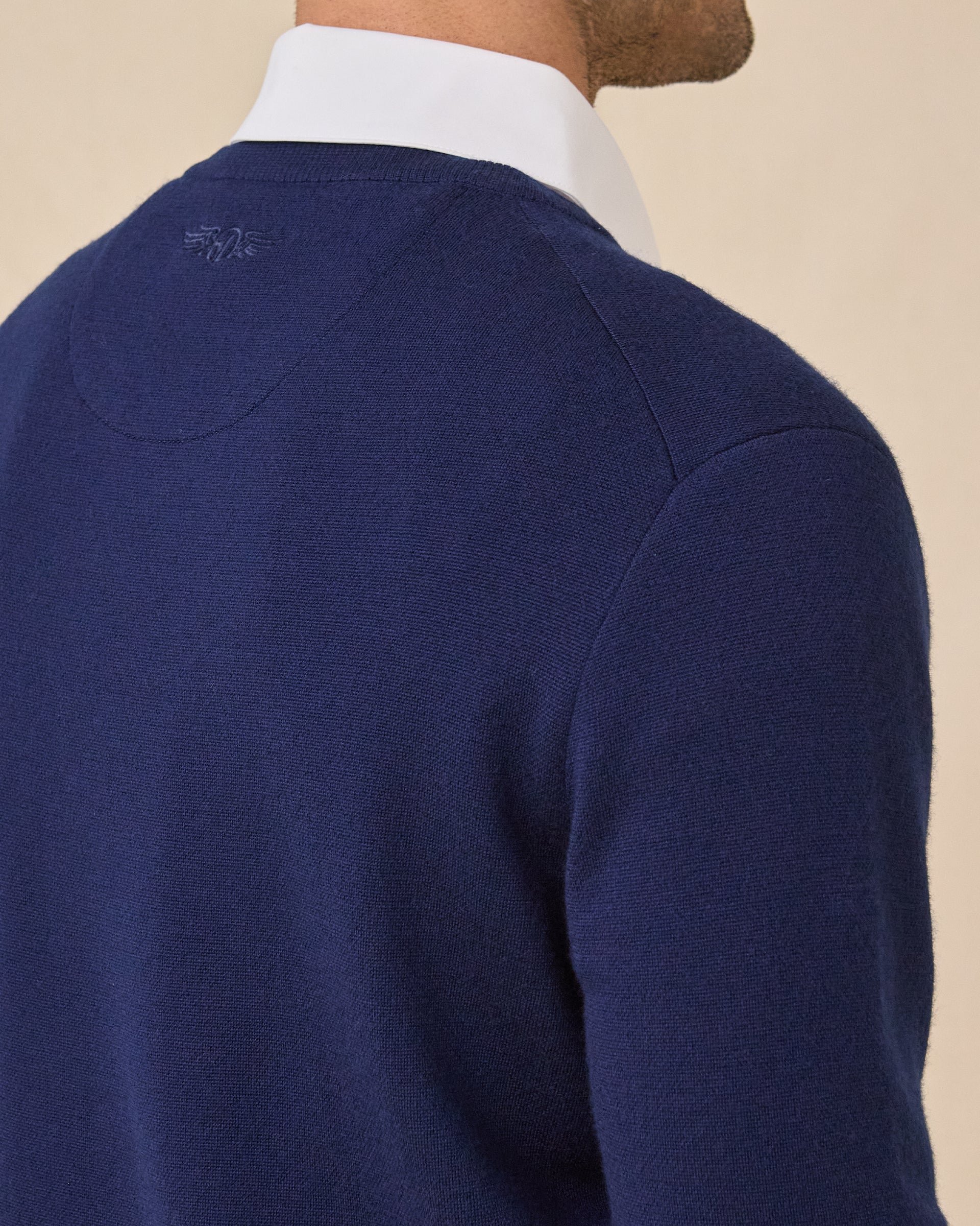Close-up back view of a model wearing a navy blue knit sweater over a white collared shirt, against a plain beige background.