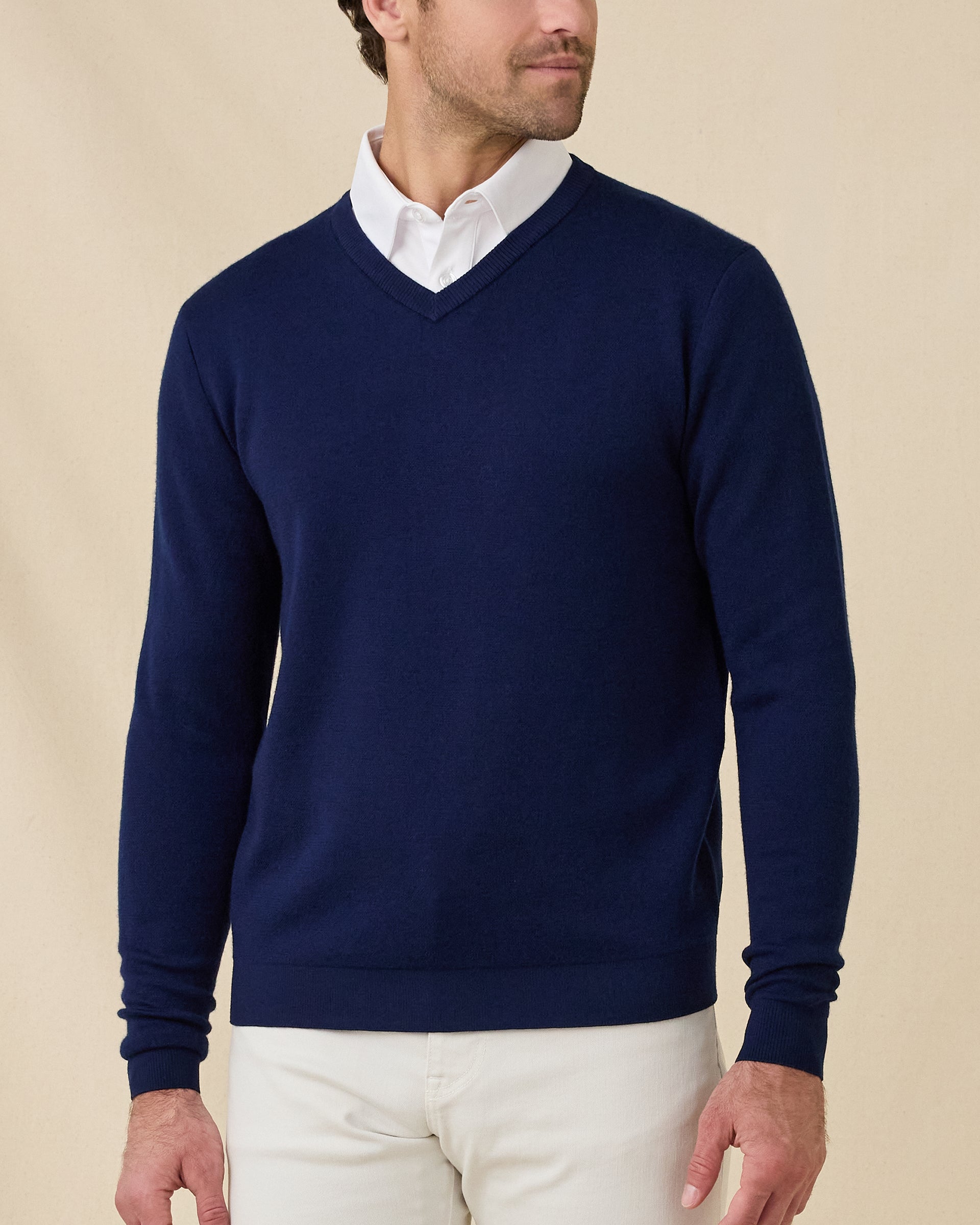 Front view of a model wearing a navy blue v-neck knit sweater over a white collared shirt and stone colored pants, photographed against a plain beige studio background.