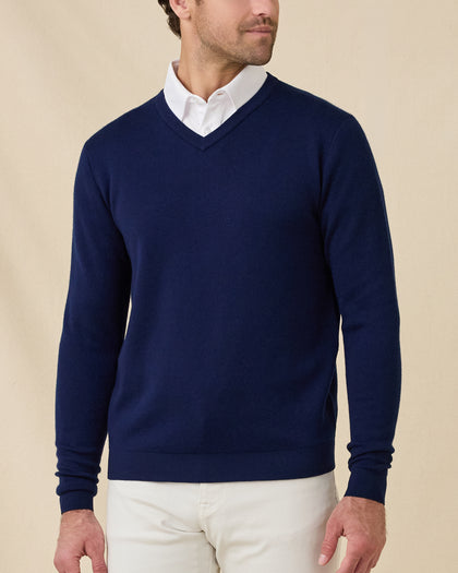 Front view of a model wearing a navy blue v-neck knit sweater over a white collared shirt and stone colored pants, photographed against a plain beige studio background.