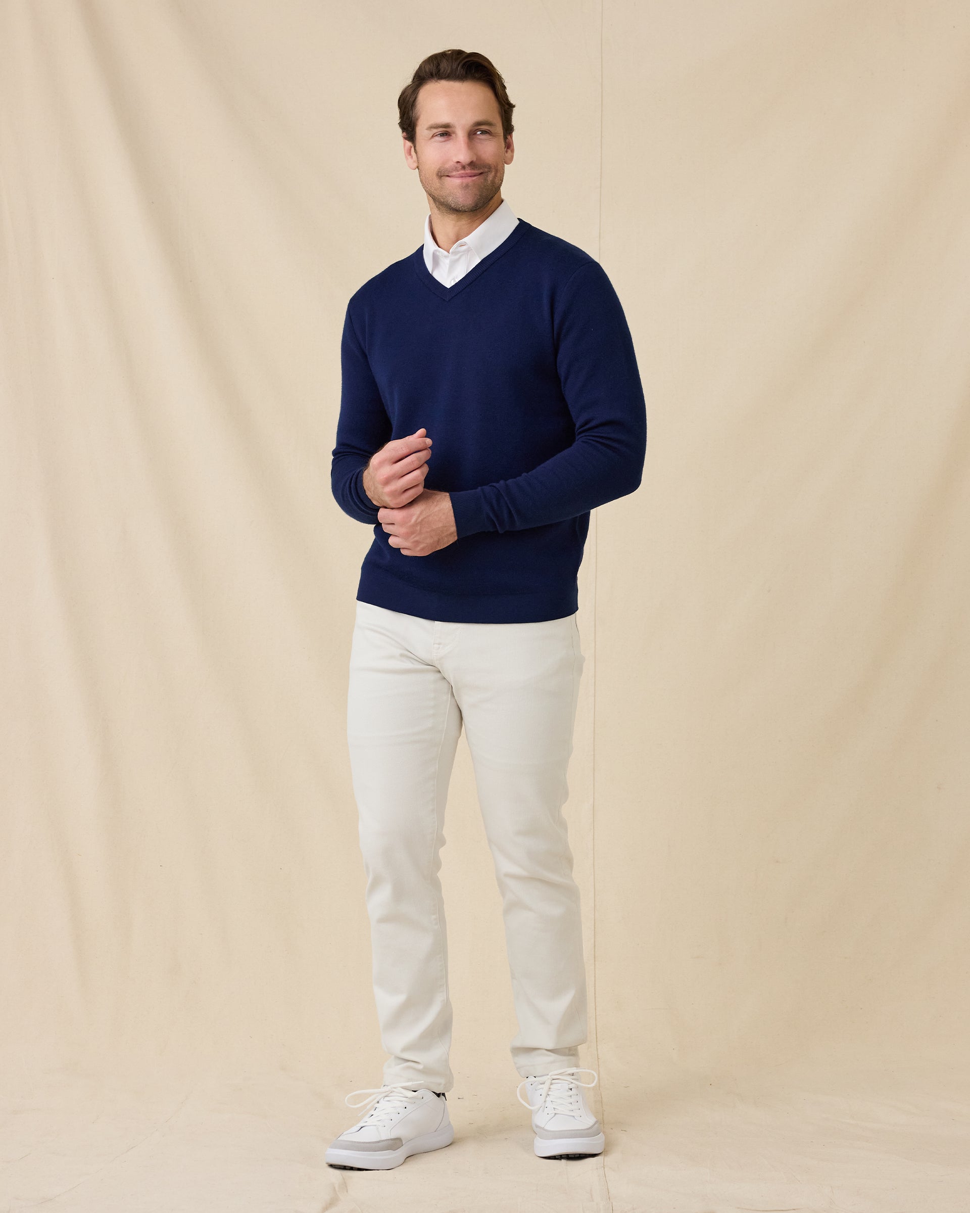 Model wearing a navy blue V-neck knit sweater over a white collared shirt, paired with stone colored pants and white sneakers, standing against a beige fabric backdrop.