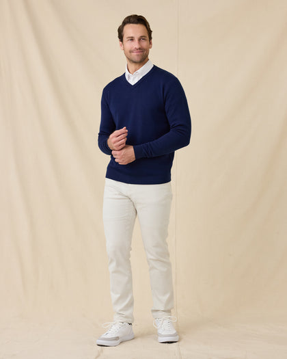 Model wearing a navy blue V-neck knit sweater over a white collared shirt, paired with stone colored pants and white sneakers, standing against a beige fabric backdrop.