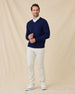 Model wearing a navy blue V-neck knit sweater over a white collared shirt, paired with stone colored pants and white sneakers, standing against a beige fabric backdrop.