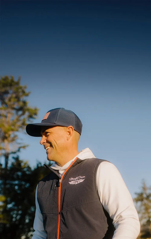 Smiling golfer wearing the Zephyr Vest in New Indigo over a White Gridlock Hoodie, paired with a Republic Hat in Navy on a sunny golf course.