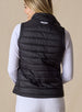 Cloud Vest in Black, back view on model featuring a black quilted vest with Straight Down embroidered logo at the center back neck styled with a white long sleeve top and white pants