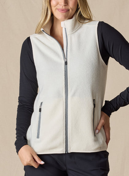 Lucia Vest in Cream, front view on model featuring an ivory fleece full-zip vest with mock neck collar, grey contrast zipper, and dual zip pockets styled with a black long sleeve top and black pants