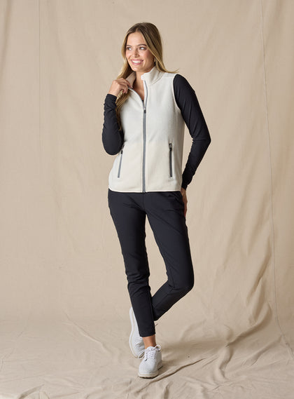 Lucia Vest in Cream, full length front view on model featuring an ivory fleece full-zip vest with mock neck collar and grey contrast zipper styled with a black long sleeve top, black pants, and white shoes