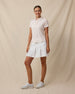 Full-length view of a woman wearing the Huli Polo in Peach with a white pleated skort and white golf shoes.