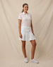 Full-length three-quarter view of a woman wearing the Huli Polo in Peach paired with a white pleated skort and white golf shoes.