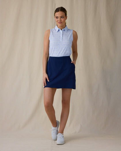 Women's Hydra Sleeveless Polo in Lavender, full-length front on-model view with a relaxed stance featuring a white sleeveless polo with an allover small floral print in light blue tones, spread collar, and two-button placket, paired with a navy skort and white shoes