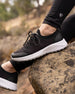 Swing Shoe in Black, lifestyle side view on model featuring the black knit upper with burgundy and purple speckle detail and white midsole styled with black leggings resting on a rock outdoors