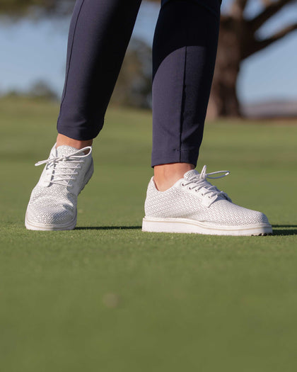 Bella Perforated Derby Shoe in White, on model view on a golf course showing both shoes worn with navy pants, featuring the all-over perforated floral cutout pattern and white platform sole