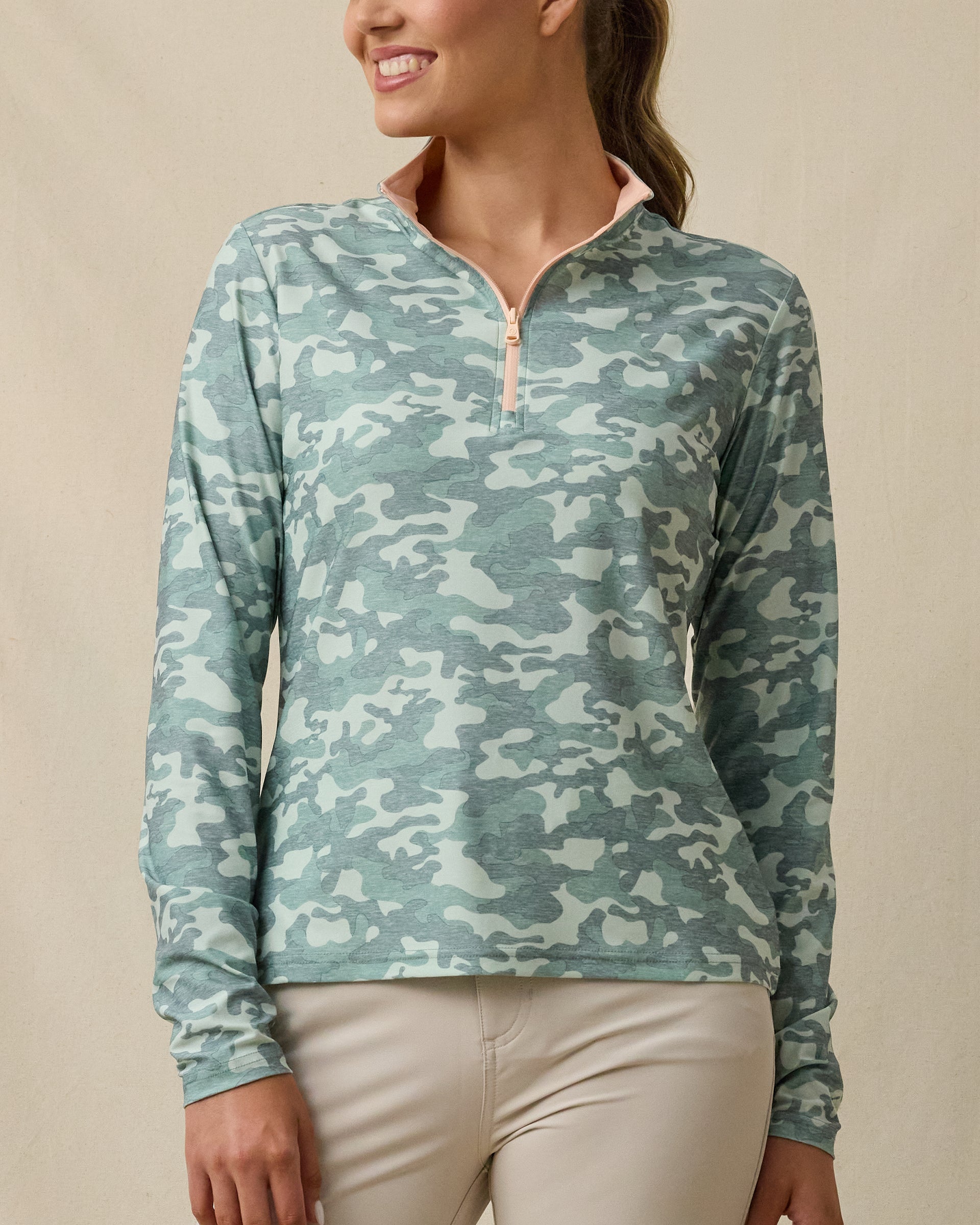 Front close-up of a woman wearing a long-sleeve quarter-zip top with a lighter and darker green camouflage print, light peach zipper, and light peach contrast collar.